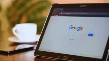 Tablet on a wooden table displaying Google’s homepage with a white cup in the blurred background, cozy cafe setting.