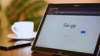 Tablet on a wooden table displaying Google’s homepage with a white cup in the blurred background, cozy cafe setting.