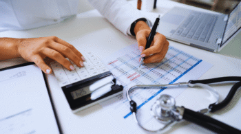 Person at a desk reviewing a chart with a calculator, pen in hand, and a laptop nearby, suggesting medical billing or financial analysis in a healthcare setting.