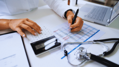 Person at a desk reviewing a chart with a calculator, pen in hand, and a laptop nearby, suggesting medical billing or financial analysis in a healthcare setting.