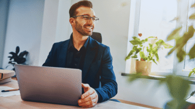 Professional man in a navy blazer using a laptop at an office desk, smiling as he looks toward the window behind him,