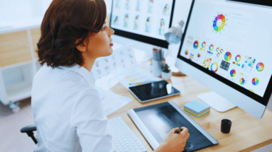 Designer at a desk using a drawing tablet with dual monitors showing color charts and graphs