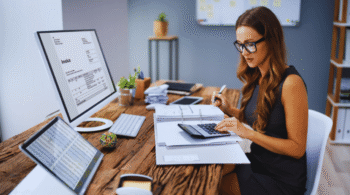Businesswoman wearing glasses reviews an invoice on a large computer monitor while counting with a calculator and organizing papers at a wooden desk.