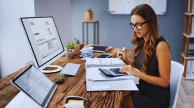 Businesswoman wearing glasses reviews an invoice on a large computer monitor while counting with a calculator and organizing papers at a wooden desk.