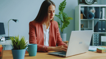 Woman in a coral blazer typing on a silver laptop at a wooden desk with a teal mug and potted plant nearby in a tidy home office.