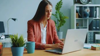 Woman in a coral blazer typing on a silver laptop at a wooden desk with a teal mug and potted plant nearby in a tidy home office.
