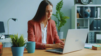 Woman in a coral blazer typing on a silver laptop at a wooden desk with a teal mug and potted plant nearby in a tidy home office.