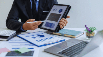 Businessperson in a suit holds a tablet displaying charts as they review reports at a desk.