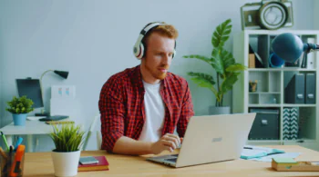 Red-haired man wearing white headphones works on a silver laptop at a wooden desk in a bright home office.